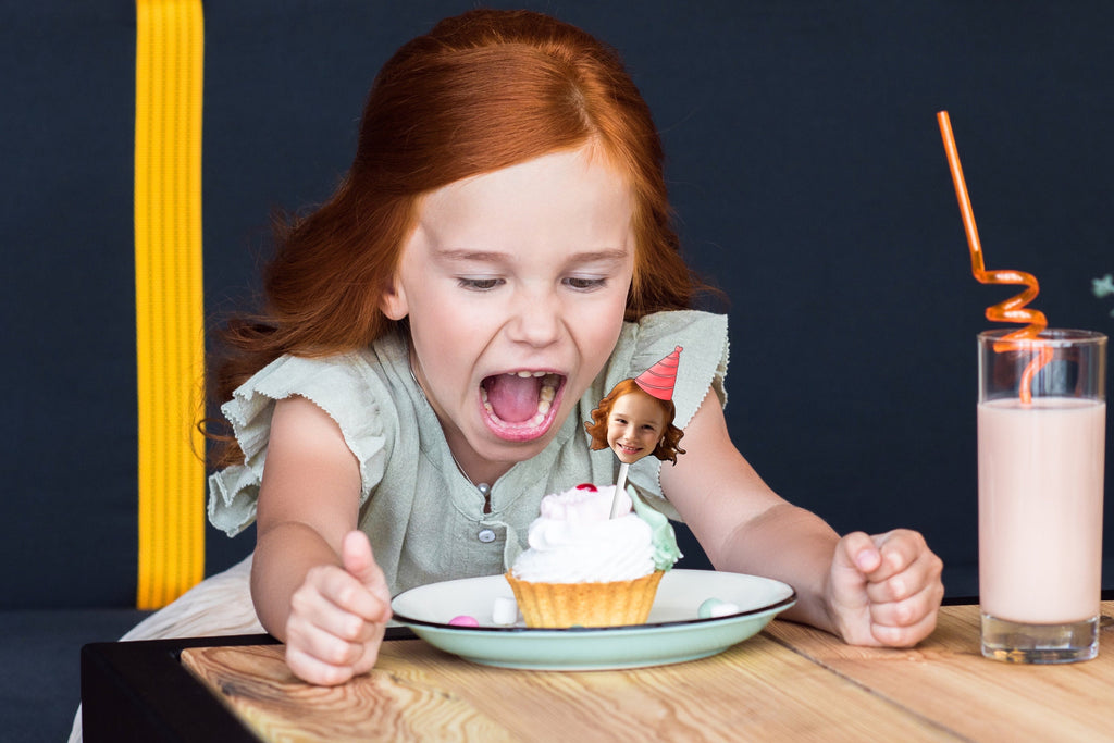 A festive decoration featuring a cut-out of a person&#39;s head wearing a colorful party hat, placed on top of a delicious cupcake, perfect for a birthday celebration.