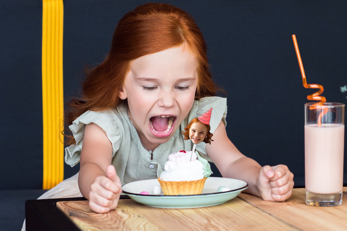 A festive decoration featuring a cut-out of a person's head wearing a colorful party hat, placed on top of a delicious cupcake, perfect for a birthday celebration.
