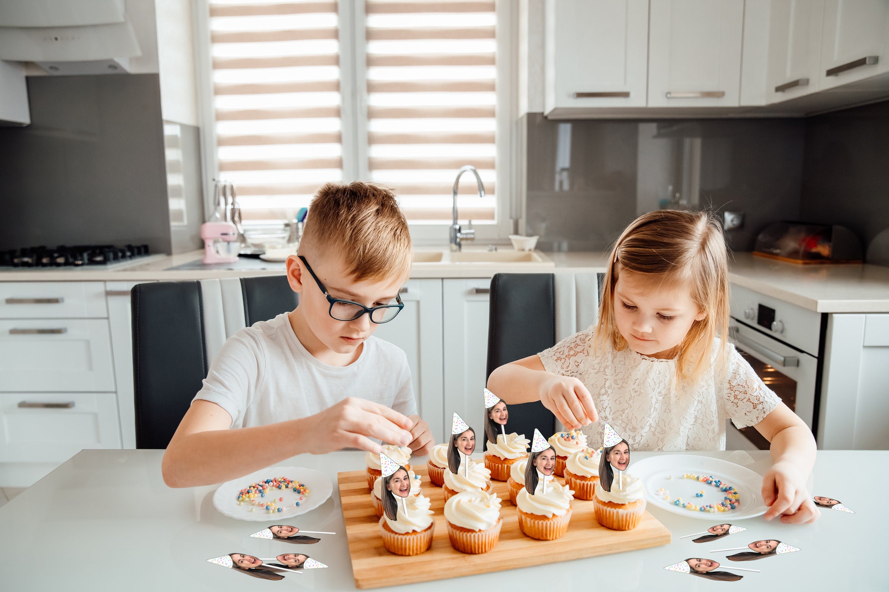 Two children joyfully decorating cupcakes with colorful sprinkles, while each cupcake is topped with a playful cake topper featuring their mum&#39;s head wearing a festive party hat, creating a fun birthday atmosphere.